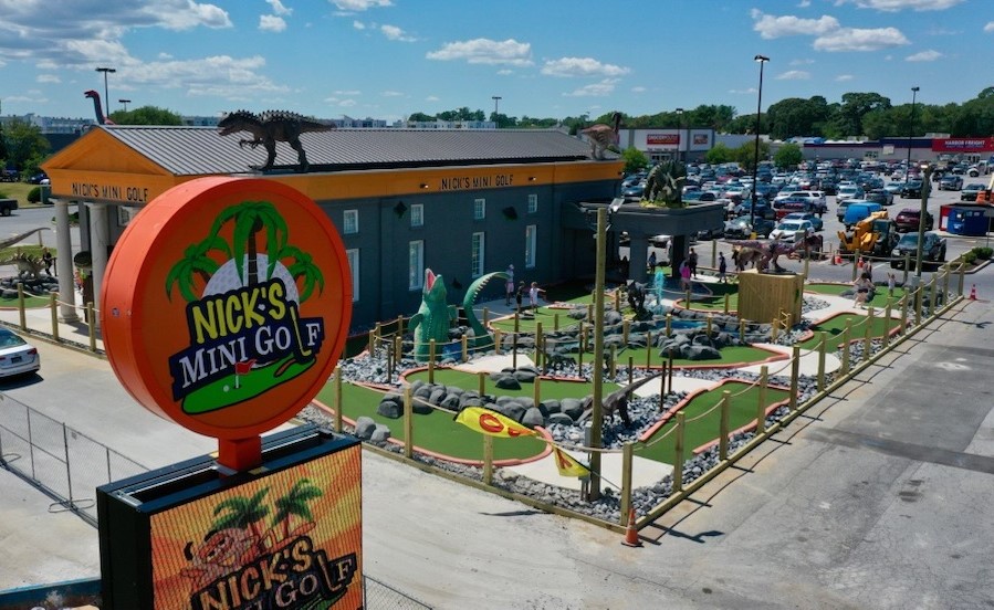 An aerial view of Nick's Mini Golf, featuring a large orange circular sign with "NICK'S MINI GOLF" text and green palm tree designs. The facility has a blue building with an orange roof topped by a dinosaur sculpture. The miniature golf course shows multiple putting greens with various obstacles, surrounded by a parking lot filled with cars. The scene is set against a blue sky with white clouds on a sunny day.